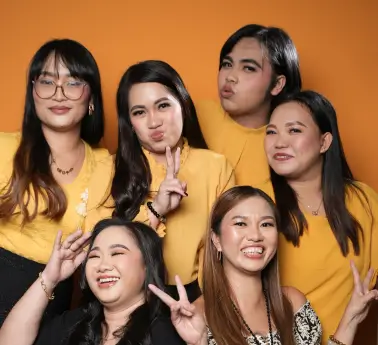 A group of women smiling and posing for a photo indoors, showcasing their friendship. They have varying hairstyles and are dressed in casual clothing against a wall. The atmosphere feels fun and lively.