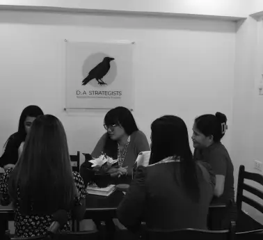 A group of women sitting at a table in an indoor setting, with a whiteboard in the background. They are dressed in various clothing styles and are engaged in conversation. The image is presented in black and white.