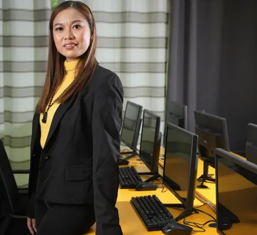 A woman standing and smiling in front of a row of computers, dressed in a jacket, inside an office building.