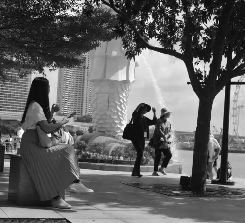 A group of people wearing various clothing styles sitting on a bench in front of a fountain, surrounded by trees. The image is in black and white.