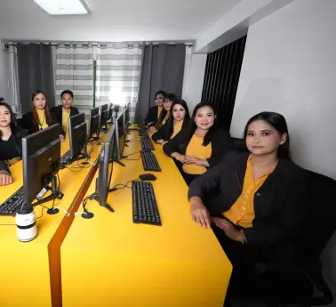 A group of women sitting at a desk with computers in an office setting, smiling and engaged in work. The room features a wall and furniture typical of an indoor workspace.