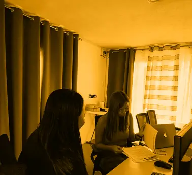 A group of women sitting at a desk in an office setting, working on computers with curtains and a window in the background.