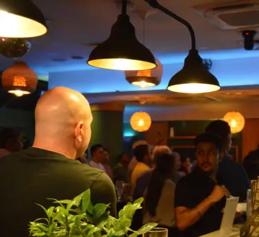 A man sitting at a table in a restaurant, surrounded by indoor plants and a bar. The ceiling and walls are visible in the background.