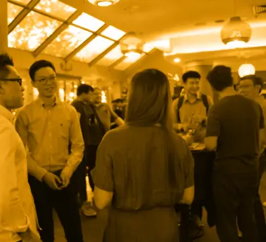 A diverse group of people standing together in a room, some wearing various types of clothing. The ceiling is visible above them, and they appear engaged in conversation or activity.