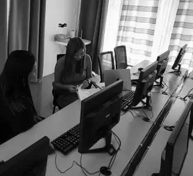 A group of people sitting at a desk in an office setting, with a personal computer on the desk. The scene is captured in black and white, showing individuals engaged in their work while sitting by a window.