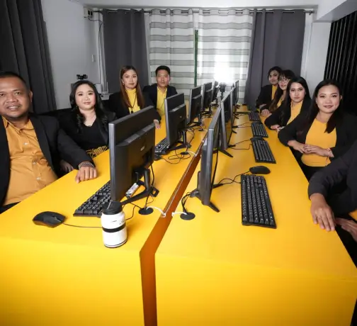 A group of people sitting at a table in an indoor office environment, each using computers. They are wearing various styles of clothing and appear to be engaged and smiling. The setting includes furniture typical of an office space, with a wall in the background.