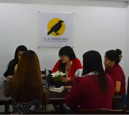 A group of women sitting at a table with a whiteboard in the background, engaged in discussion. The setting is indoors, featuring furniture related to education and learning.