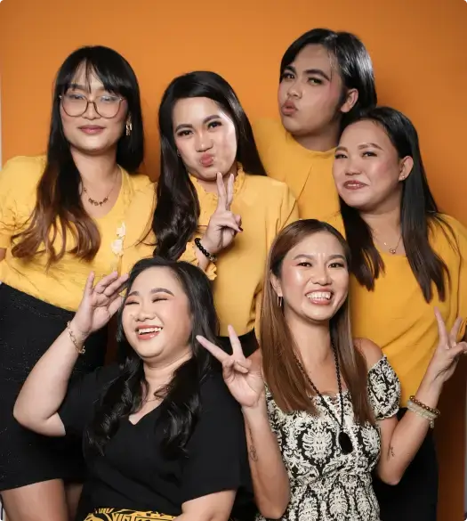 A group of women smiling and posing for a picture indoors, standing close together in a friendly manner. They are dressed in fashionable clothing and have varying hairstyles. The background features a wall that adds to the casual atmosphere of the gathering.