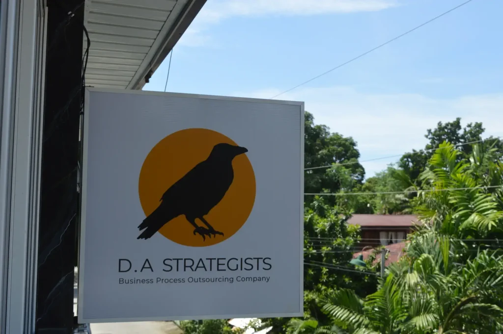 A sign on a building that reads "D.A STRATEGISTS Business Process Outsourcing Company," surrounded by outdoor elements such as a tree and a blue sky. A bird is perched nearby. The sign is yellow and features text.